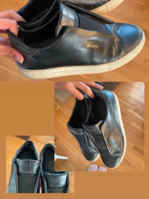 Svarta sneakers från Axel Arigato - Säljer ett par svarta sneakers från Axel Arigato med vit sula och diskret logga på sidan. Skorna har en clean slip-on design med elastiska paneler och är tillverkade i läder. Perfekt för dig som gillar minimalistisk stil. Släpppt lite i tyget inuti vid hälen bak. Men inget som stör eller känns eller syns! :) 