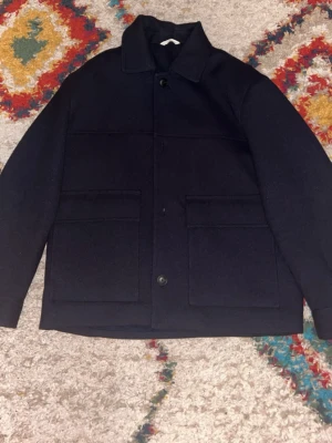 Mörkblå overshirt med stora fickor - Stilren mörkblå overshirt med klassisk krage och stora fyrkantiga fickor framtill. Rak passform och knäppning med knappar. Materialet ser ut att vara tjockt och kan vara ullblandning, perfekt för lager på lager. Enkel och clean design utan synliga loggor.