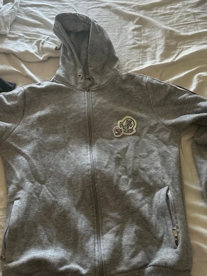 Grå Moncler hoodie med dragkedja - Säljer en grå hoodie från Moncler med dragkedja och huva. Den har ett Moncler-märke på bröstet och röda/vita detaljer längs axlarna. Två fickor med dragkedja framtill. Perfekt för dig som gillar stilrena och exklusiva plagg.