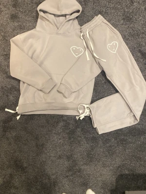 Carsicko tracksuit - Mysigt grått loungewear-set från Carsicko med hoodie och matchande byxor. Båda delarna har vita dragskor och ett broderat hjärta med smiley på bröstet och benet. Tillverkat i mjukt fleecematerial, perfekt för chill dagar hemma. Du får med både hoodie och byxor