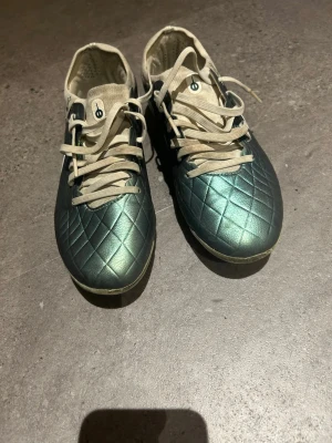 Nike fotbollsskor med metallic färg - Säljer ett par grymma fotbollsskor från Nike med metallicgrön ovandel och vita detaljer. Skorna har quiltad struktur på ovandelen och vita snören samt innersida. Dem är som vanligt små i storleken så snarare storlek 36,5/37.