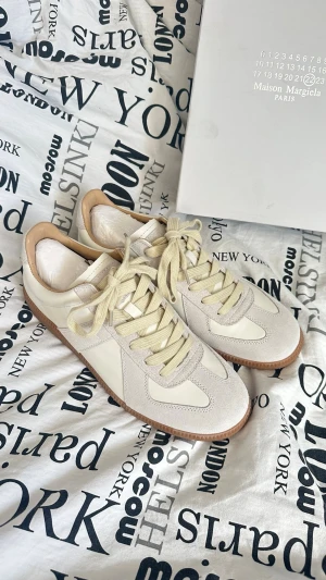 Maison Margiela vita sneakers - Maison Margiela sneakers i vitt med beige detaljer och gummisula. Skorna har klassisk låg profil, beige snörning och kombinerar läder och mocka för en stilren look. Perfekt för dig som vill ha något exklusivt och trendigt.