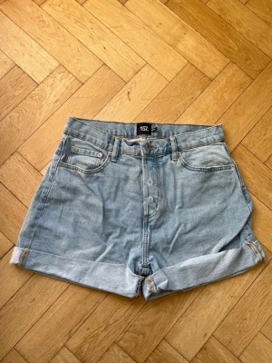 Ljusblå denimshorts från Lager 157 - Säljer ett par ljusblå denimshorts från Lager 157 med klassisk femficksdesign och uppvikta benslut. Shortsen har hög midja och bälteshällor, perfekta för sommardagar. Stängs med knapp och dragkedja framtill. I modellen Diba.