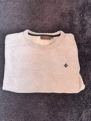 Ljusgrå sweatshirt från Morris - Ljusgrå sweatshirt från Morris med rund hals och diskret svart logotyp broderad på bröstet. Tröjan har ribbade muddar och en patch nertill på framsidan. Perfekt för en avslappnad och stilren look.