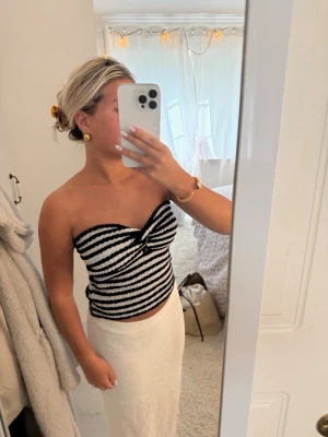 Randig bandeau tubtopp i svart och vitt - Säljer en snygg bandeau tubtopp med svartvita ränder och twistad detalj framtill. Toppen är axelbandslös och har en croppad passform som passar perfekt till höga kjolar eller byxor. Materialet känns mjukt och stretchigt, troligen bomull eller syntet.