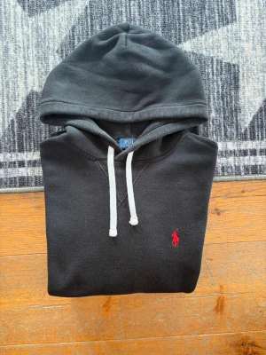 Svart Polo Ralph Lauren Hoodie - Storlek M - Hej! 👋 Säljer en klassisk svart Polo Ralph Lauren hoodie i storlek M. Den har den ikoniska röda pony-loggan broderad på bröstet 🐎 Tröjan är i bra skick, djupsvart i färgen (inte solblekt) och inte särskilt nopprig ✨ Den har den sköna fleece-insidan kvar och är 100% äkta med alla tvättlappar kvar ✅ Ingen QR-kod på denna modell. Ett tidlöst premium-basplagg som passar till allt och håller stilen år efter år 👕 Fraktar snabbt! 🚛📦💬💯 