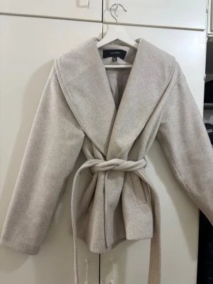 Beige kappa med knytskärp från Vera moda - Stilren beige kappa från Vera Moda med bred krage och knytskärp i midjan. Kappan har raka ärmar, två fickor framtill och är tillverkad i ett mjukt, ull-liknande material. Perfekt för dig som gillar minimalistisk och trendig stil.