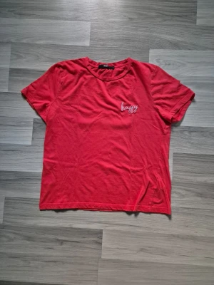 Röd t-shirt med broderad text - Röd t-shirt från Bik Bok med korta ärmar och rund halsringning. På bröstet finns en liten broderad vit text där det står 'happy'. Tillverkad i mjuk bomull för en skön känsla. Perfekt för en avslappnad och enkel stil.