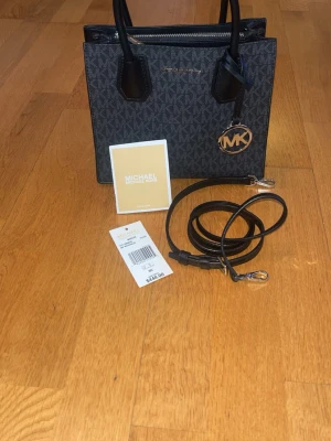 Svart handväska från Michael Kors (helt ny) - Snygg svart handväska från Michael Kors med klassiskt MK-mönster och guldfärgad logga. Väskan har två handtag, avtagbar axelrem och dragkedja upptill. Tillverkad i tåligt konstläder och har detaljer i svart och guld. Perfekt för dig som gillar stilrena accessoarer. 100% äkta, nytt skick. 