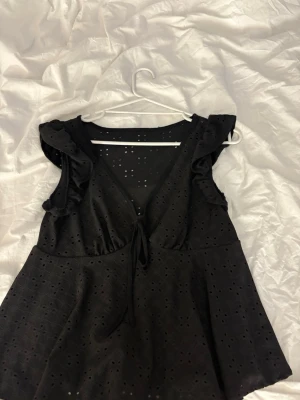 Svart blus med volang och knyt - Snygg svart blus med volangdetaljer vid axlarna och v-ringning med knyt framtill. Blusen har ett broderat hålmönster som ger en luftig känsla och är ärmlös. Perfekt för dig som vill ha en feminin och trendig topp.