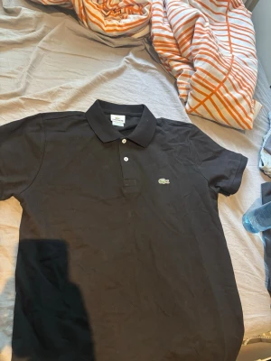 Svart pikétröja från Lacoste - Klassisk svart pikétröja från Lacoste med två vita knappar och den ikoniska krokodilloggan på bröstet. Tröjan har krage och korta ärmar, perfekt för en clean och stilren look. Materialet är bomull och passformen är normal.