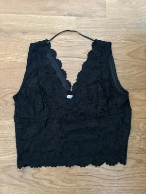 Svart spetsig croppad topp från Amisu - Svart croppad topp från Amisu med v-ringad hals och vacker spets över hela plagget. Ärmlös design och dekorativa kanter som ger en elegant look. Perfekt för att lyfta din outfit med lite extra edge.