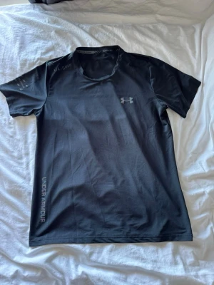 Svart tränings-tshirt från Under Armour - Säljer en svart tränings-tshirt från Under Armour med logga på bröstet och text på ärmen. Materialet är lätt och snabbtorkande, perfekt för intensiva pass. Rund hals och korta ärmar ger en bekväm passform. Snygg och funktionell för gym eller löpning.