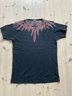 Svart t-shirt från Marcelo Burlon  - Svart t-shirt med korta ärmar och en unik röd fjäderliknande detalj runt halsringningen. 