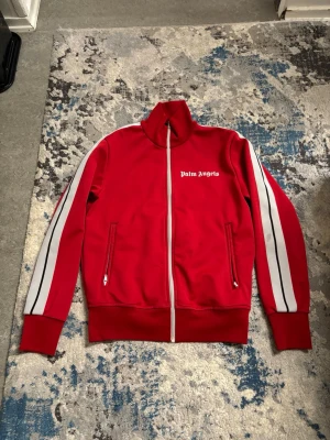 Röd zip-tröja från Palm Angels - Palm Angels röd zip-tröja med vita och svarta ränder längs ärmarna. Tröjan har hög krage, hel dragkedja och två fickor med dragkedja. Logotypen är tryckt på bröstet. Perfekt för en sportig och trendig look.