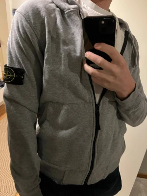 Grå zip hoodie från Stone Island - Säljer en grå hoodie från Stone Island med dragkedja framtill och klassisk logopatch på vänster ärm. Hoodien har huva med dragsko och två fickor fram. Tillverkad i mjukt sweatshirtmaterial, perfekt för en avslappnad stil.
