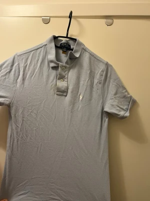 Ljusblå pikétröja från Polo Ralph Lauren - Klassisk ljusblå pikétröja från Polo Ralph Lauren med korta ärmar och krage. Tröjan har två knappar framtill och den ikoniska broderade loggan på bröstet. Perfekt till jeans eller shorts för en clean och stilren look. Tröjan har en liten gul fläck som ni ser på tredje bilden