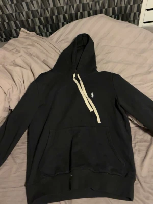 Svart hoodie från Polo Ralph Lauren - Snygg svart hoodie från Polo Ralph Lauren med vit dragsko och klassisk logga broderad på bröstet. Hoodien har stor huva och magficka framtill. Tillverkad i mjuk bomullsblandning som är perfekt för chill dagar.