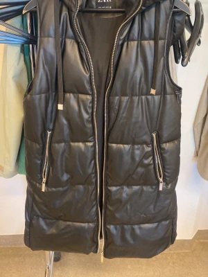 Svart lång dunväst från Zara - Snygg svart lång dunväst från Zara med glansig finish och huva. Västen har två dragkedjefickor framtill, breda kanaler och dragsko vid huvan. Perfekt för lager-på-lager och streetstyle. Materialet är quiltat och ger en modern look.