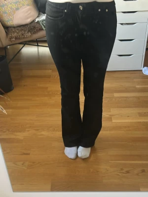Svarta bootcut jeans med fickdetaljer - Säljer ett par svarta bootcut jeans med snygga fickor bak som har lock och silverfärgade knappar. Jeansen har klassisk femficksdesign och bälteshällor. Perfekta för dig som gillar en tidlös och clean look.