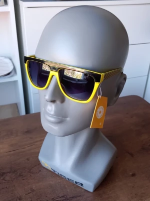 Gula solglasögon från Sunshine  - Coola solglasögon från Sunshine med gul båge och svarta detaljer. Sidorna har ett randigt mönster som ger en extra touch. Glasen är mörka och bågarna är i syntetmaterial. Perfekta för att sticka ut i sommar!