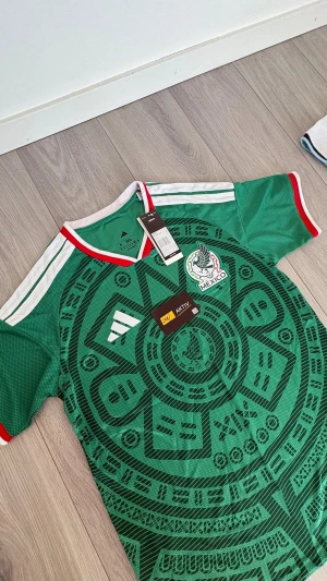 Mexikansk landslagströja Adidas grön - Grön Mexiko fotbollströja från Adidas med svart aztekiskt mönster, vita axelränder och röd-vit krage. Broderad Mexiko-logga på bröstet och klassisk sportig passform. Perfekt för fotboll eller träning.