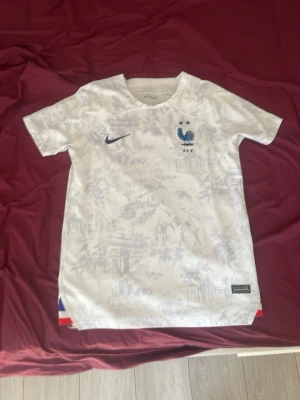 Frankrike fotbollströja Nike vit - Snygg vit Frankrike fotbollströja från Nike med diskret mönster, blå Nike-logga och franska landslagets emblem på bröstet. Korta ärmar och rund hals. Tricolore-detaljer i nederkant. Perfekt för fotbollsfans!