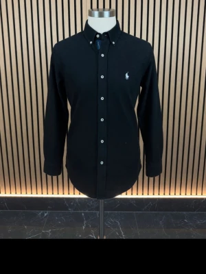 Svart skjorta från Ralph Lauren - Klassisk svart skjorta från Ralph Lauren med vit broderad logga på bröstet. Skjortan har knappar framtill, button-down krage och långa ärmar. Perfekt för en stilren look.