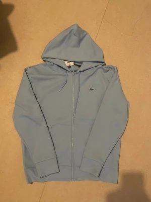 Ljusblå hoodie från Lacoste - Snygg ljusblå hoodie från Lacoste med dragkedja framtill och klassisk krokodillogga på bröstet. Hoodien har huva med dragsko och två fickor framtill. Tillverkad i mjukt bomullsmaterial som är skönt att ha på sig.