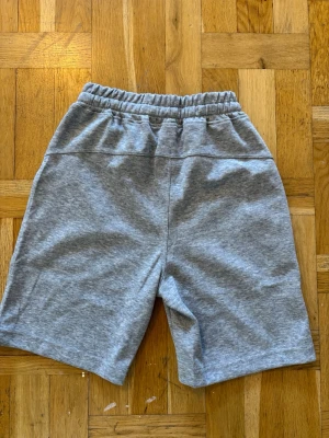 Grå Essentials Fear of God shorts - Snygga grå sweatshorts från Essentials Fear of God med elastisk midja och snörning. Shortsen har två sidofickor och logga tryckt på vänster ben. Perfekta för chill dagar och streetwear vibes.