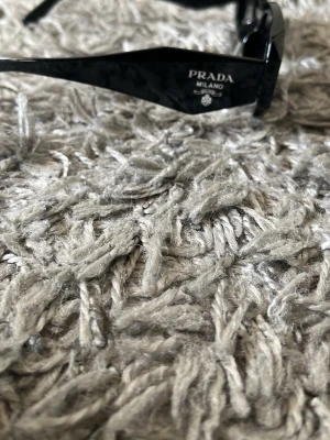 Svarta solglasögon från Prada - Snygga svarta solglasögon från Prada med rektangulära bågar och diskret logga på sidan. Glasögonen har en stilren och modern design som passar perfekt till en trendig look. Tillverkade i blankt syntetmaterial.