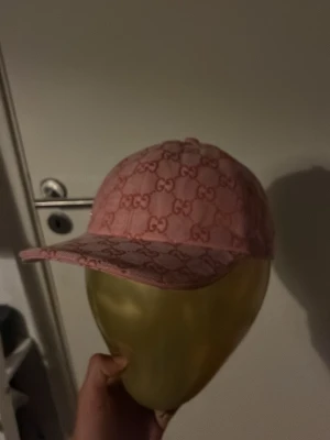 Rosa Gucci keps med GG-mönster - Säljer en snygg rosa keps från Gucci med det klassiska GG-monogrammet över hela kepsen. Kepsen har böjd skärm och justerbar rem bak. Materialet är en blandning av bomull och canvas, vilket ger en exklusiv känsla och look.