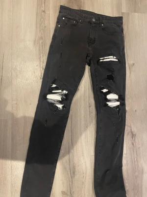 Amiri  - Säljer ett par svarta skinny jeans med stora slitningar och hål på båda benen. Jeansen har fem fickor, knappgylf och är i stretchigt material för skön passform. Perfekta för en edgy och trendig look.