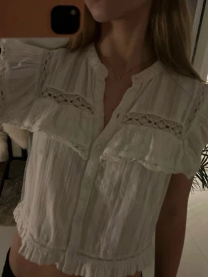 Vit blus med volanger från Zara - Söt vit blus från Zara med korta ärmar, volangdetaljer och spetsinslag. Blusen har knäppning framtill, lätt rynkade kanter och en luftig känsla.