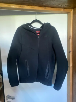 Nya Nike Tech Fleece - Svart hoodie från Nike med hel dragkedja framtill och två fickor med dragkedja. Klassisk Nike-logga på bröstet och huva. Perfekt för en avslappnad streetstyle-look. Mjuk och bekväm bomullsblandning.