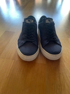 Marinblåa sneakers från J.Lindeberg - Stilrena svarta sneakers från J.Lindeberg med vit sula och svart tygöverdel. Klassisk låg modell med svarta skosnören och logga på plösen. Perfekta för en clean och avslappnad look.