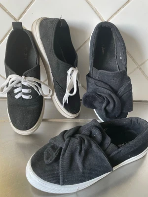 Svarta sneakers med rosett och snörning - Två par svarta låga sneakers, ett par med vita snören och ett par med bred rosett framtill. Båda har vit sula och är i tygmaterial. Perfekta för en avslappnad och trendig look.