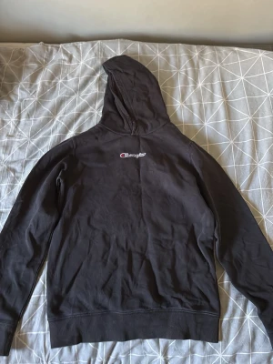 Svart hoodie från Champion - Säljer en svart hoodie från Champion med klassisk logga broderad på bröstet. Hoodien har huva och ribbade muddar vid ärmslut och nederkant. Perfekt för dig som gillar en enkel och stilren look. Materialet känns mjukt och bekvämt.