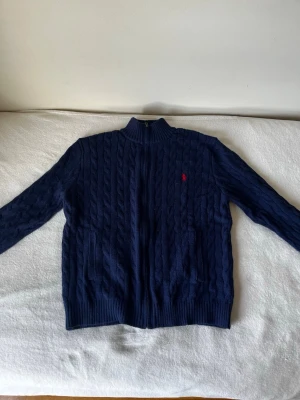 Mörkblå kabelstickad kofta Polo Ralph Lauren - Mörkblå kabelstickad kofta från Polo Ralph Lauren med dragkedja framtill, två fickor och röd broderad logga på bröstet. Hög krage och ribbade muddar. Perfekt för lager på lager och riktigt snygg till jeans.