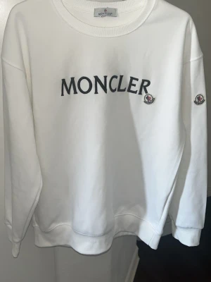 Vit Moncler sweatshirt med logga - Snygg vit sweatshirt från Moncler med stor svart logotyp på bröstet och broderad Moncler-patch på ärmen. Klassisk rund halsringning och ribbade muddar. Perfekt för en clean och stilren look.