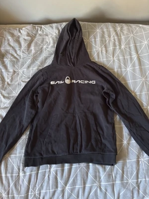 Svart hoodie från Sail Racing - Svart hoodie från Sail Racing med vit logga tryckt på bröstet. Klassisk modell med huva och känguruficka framtill. Tillverkad i mjukt bomullsmaterial och har ribbade muddar vid ärmslut och nederkant. Perfekt för en avslappnad och sportig stil.