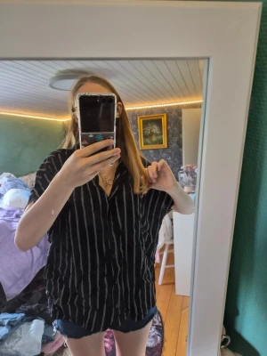 Svart randig skjorta från xlnt - Säljer en svart skjorta från xlnt med vita vertikala ränder. Skjortan har korta ärmar, knappar framtill och en loose passform. Perfekt för dig som gillar en avslappnad stil.