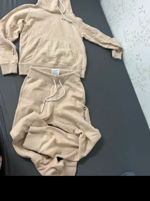 Beige loungewear set från Champion - Mysigt beige loungewear set från Champion med hoodie och matchande byxor. Hoodien har magficka, dragsko och broderad logga på bröstet och ärmen. Byxorna har resår i midjan, dragsko och logga på benet. Perfekt för chill dagar hemma.