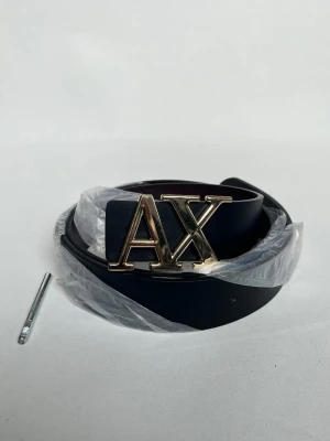 Svart skärp från Armani Exchange - Stilrent svart skärp från Armani Exchange med ett stort guldfärgat AX-spänne. Skärpet är tillverkat i skinn och har en klassisk, slät design som passar till många olika outfits.