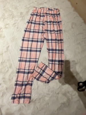 Rosa rutiga mjukisbyxor XS - Säljer ett par rosa mjukisbyxor med blått och vitt rutmönster. Byxorna har resår i midjan och är gjorda i mjukt material med 95% polyester och 5% elastan. Perfekta för chill dagar hemma eller sleepovers.
