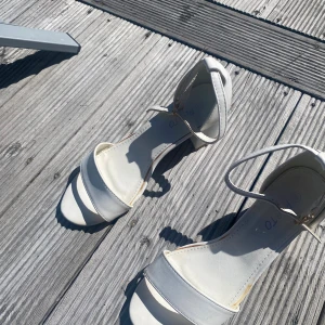 Vita sandaler med låg klack - Snygga vita sandaler med öppen tå och låg klack. De har en enkel rem över tårna och en justerbar rem runt ankeln. Perfekta för sommaren och ger en clean look till alla outfits. Tillverkade i syntetmaterial.
