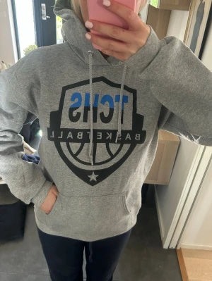 Grå TC HS Basketball hoodie - Snygg grå hoodie med TC HS Basketball-tryck i svart och blått på bröstet. Klassisk känguruficka framtill och justerbar huva med dragsko. Perfekt för dig som gillar sportig stil och vill ha något bekvämt och avslappnat. Mjuk bomullsblandning.