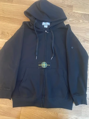 Svart hoodie från Stone Island - Svart hoodie från Stone Island med dragkedja, huva med dragsko och klassisk logotyp-patch på bröstet. Mjuk bomullskänsla och känguruficka framtill. Perfekt för en avslappnad streetwear-look.