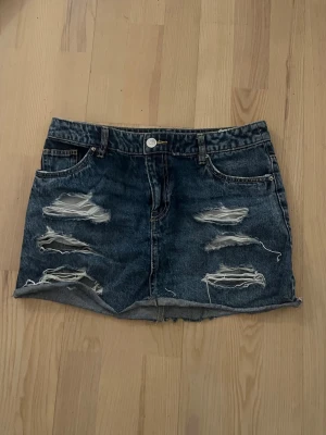 Blå jeanskjol med slitningar från H&M - Säljer en blå jeanskjol från H&M i storlek 36. Kjolen är kort och har flera slitningar framtill för en trendig, avslappnad look. Klassisk femficksmodell med knapp och dragkedja. Perfekt till sneakers eller boots.