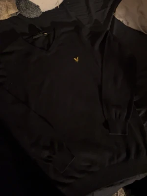 Svart stickad tröja från Lyle & Scott - Svart stickad tröja från Lyle & Scott med V-ringning och klassisk gul logga broderad på bröstet. Tröjan har långa ärmar och ribbade muddar vid ärmslut och nederkant. Perfekt för en stilren och enkel look.